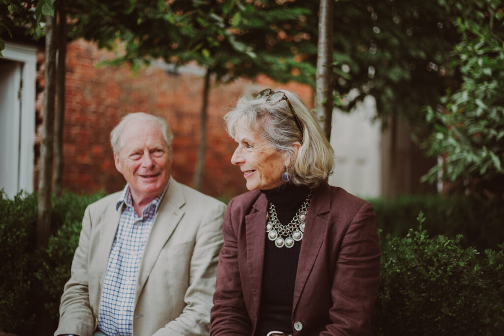 A man and lady sit next to one another outside underneath a pleached tree