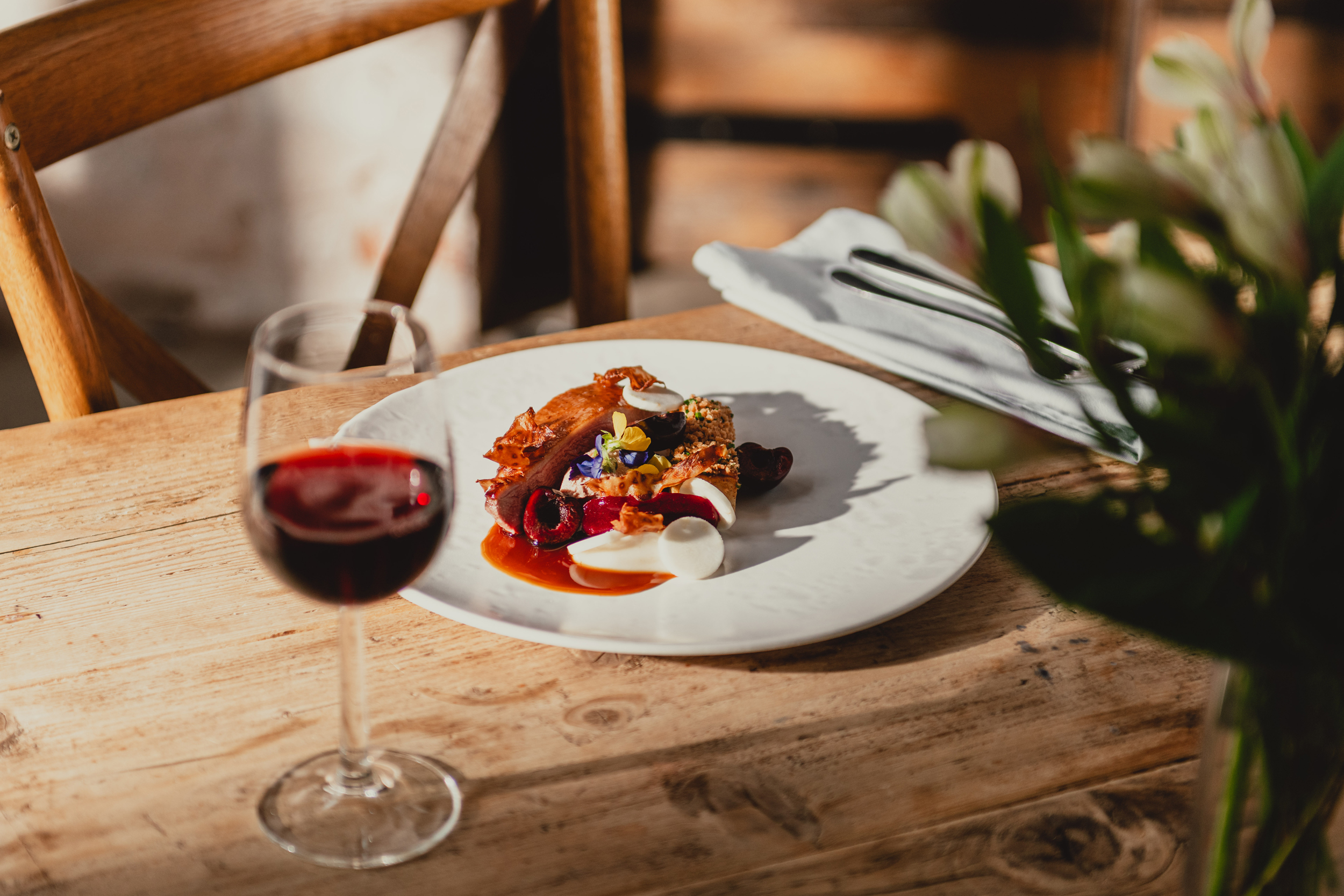 A glass of red wine on a rustic wooden table accompanies a white plate on which an elegant arrangement of meat garnished with flavourings resides.