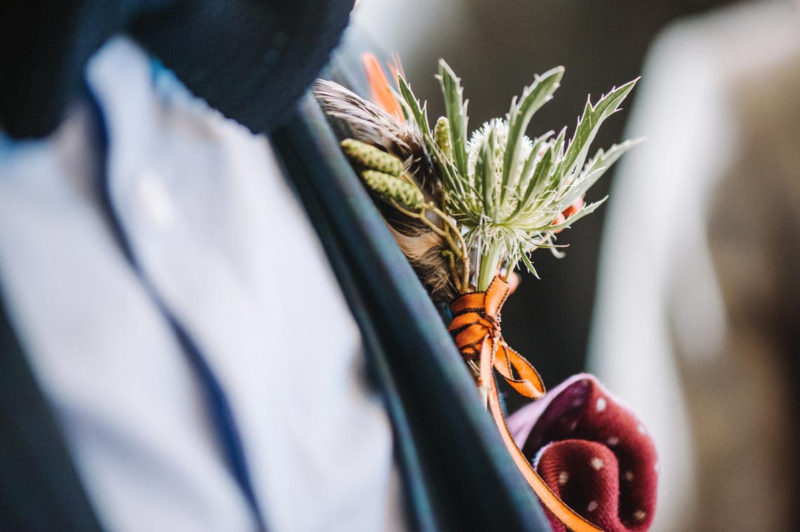 A close up of a groom's button hole tied with an orange ribbon and filled with catkins and a thistle