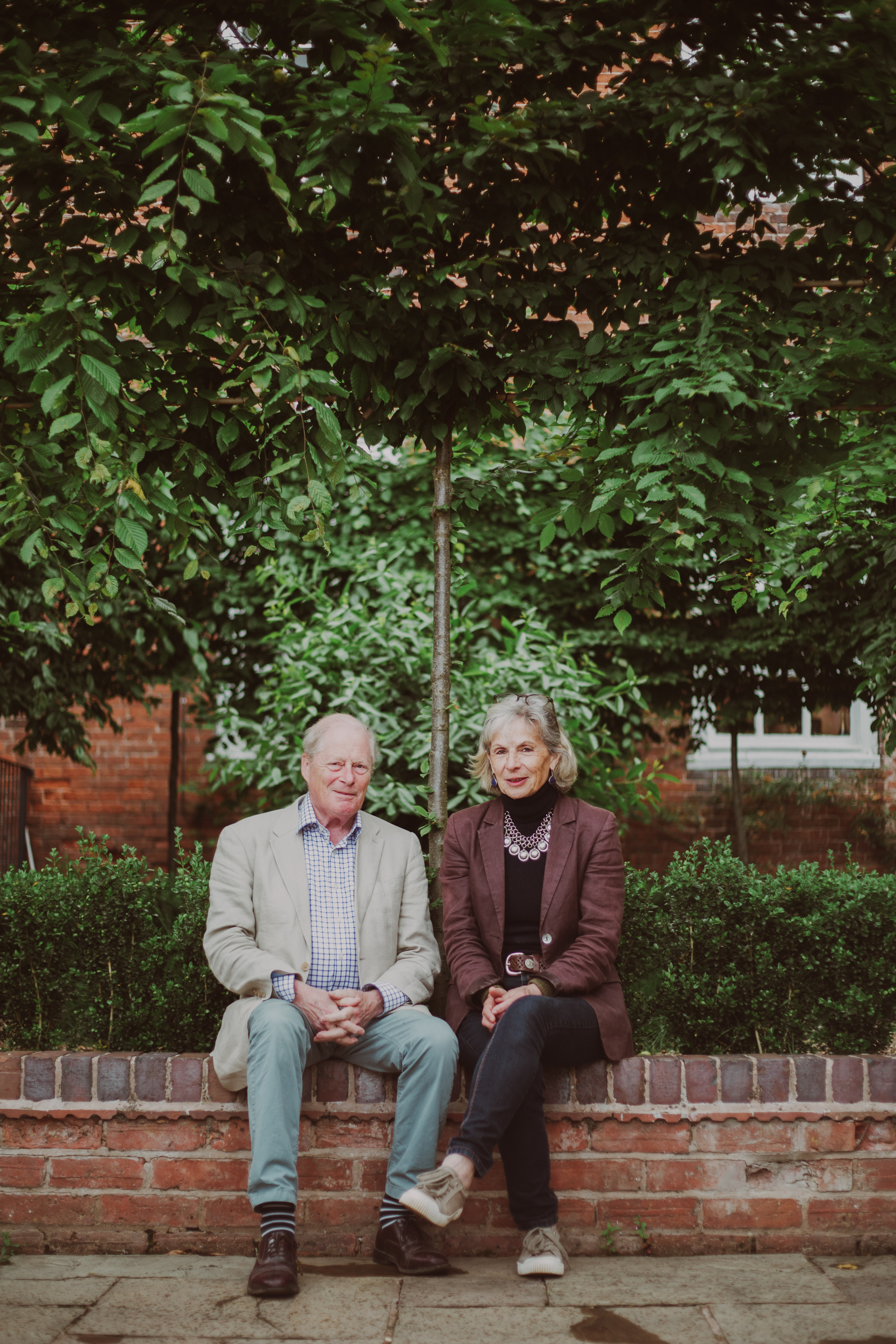 A man and lady sit next to one another outside underneath a pleached tree