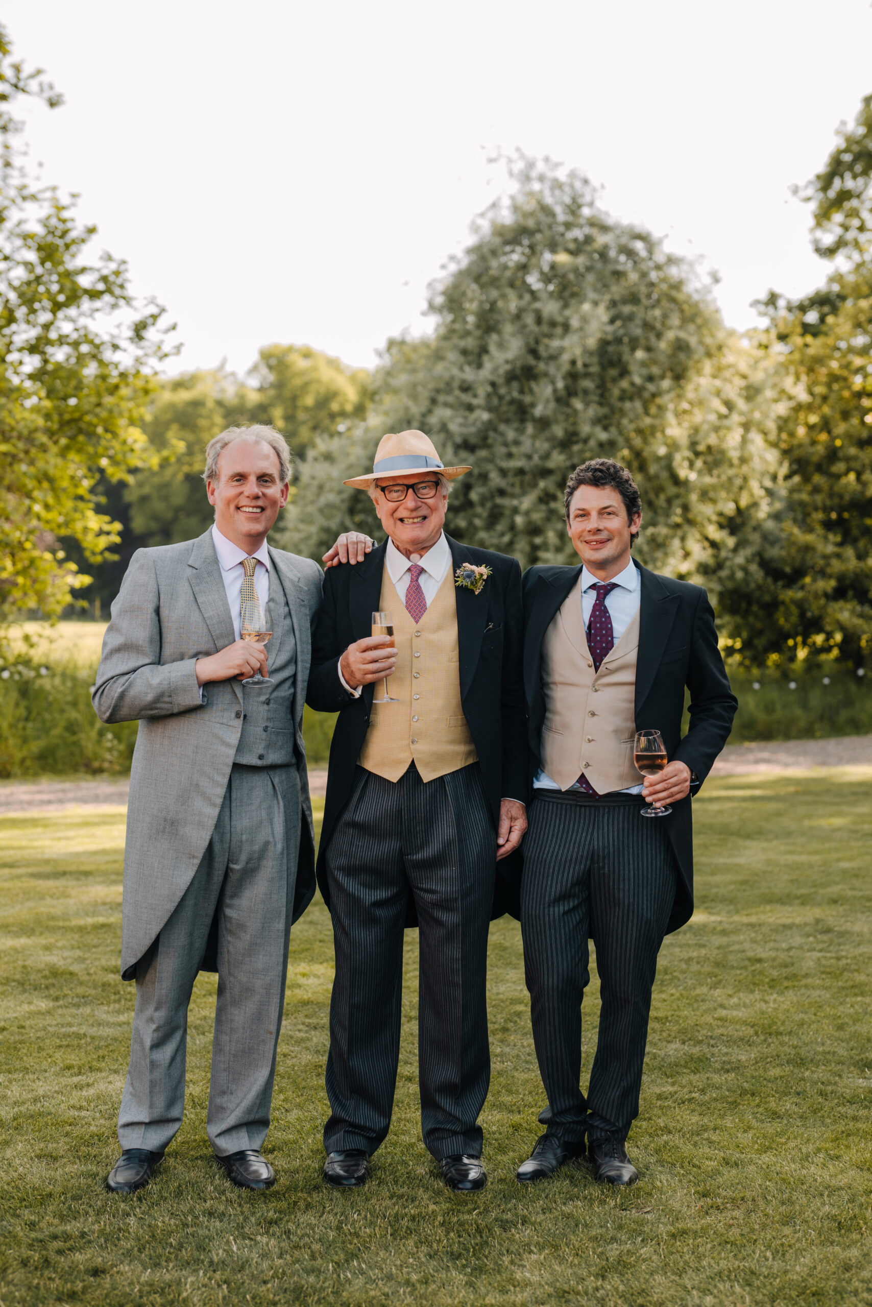 Three men wearing morning suits stand outside on a manicured lawn with mature trees in the background posing for the camera.