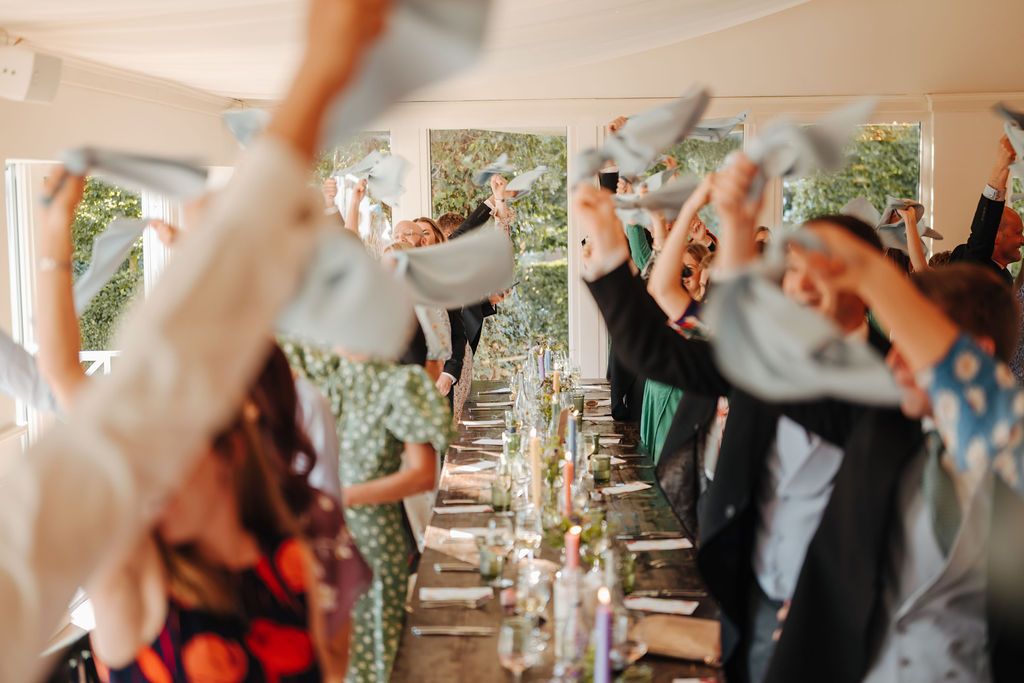 Guests at a wedding reception stand by their seats at a trestle table waving blue linen napkins