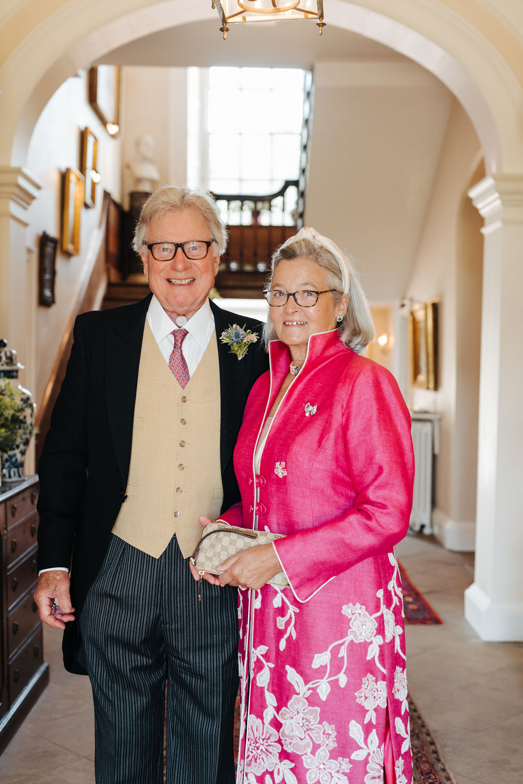 A man wearing a morning suit stands next to his wife who is wearing a pink floral outfit dressed for a wedding with an antique wooden staircase in the background behind them.