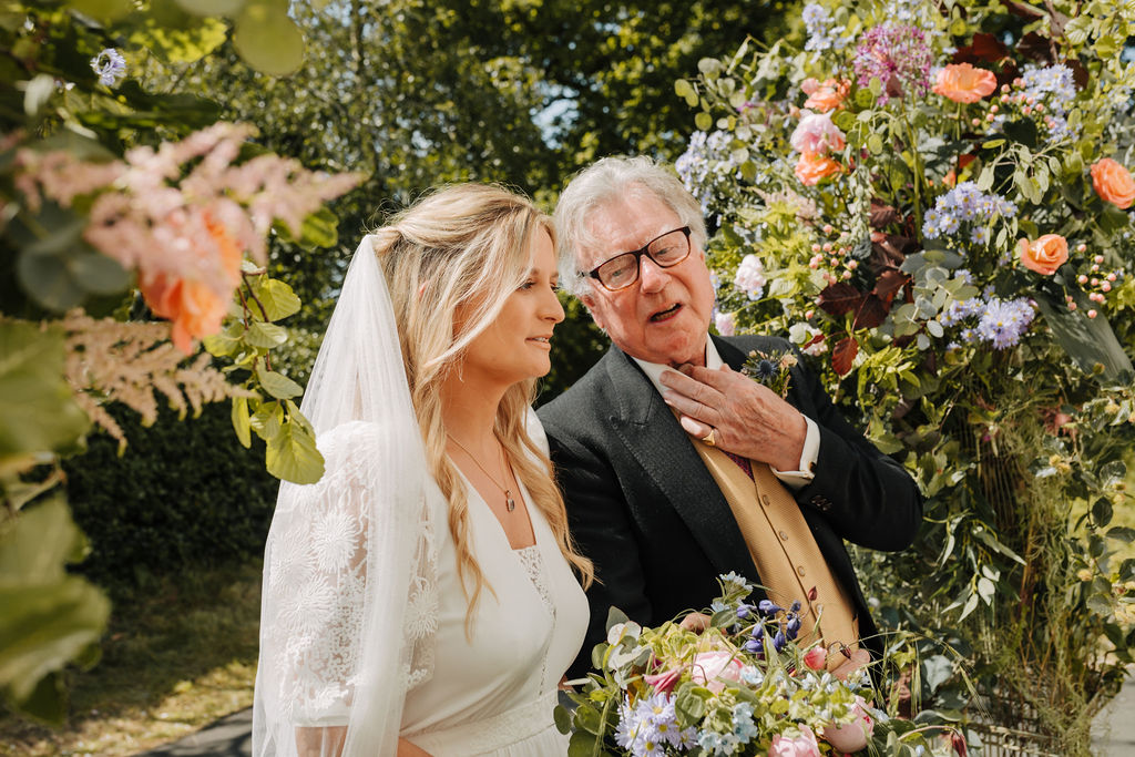 A bride and her father stand waiting to enter a church surrounded by lots of June flowers