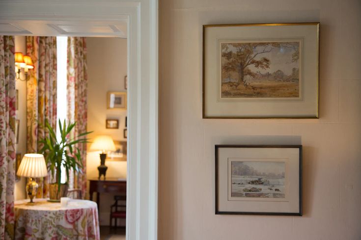 A focus on two watercolour paintings hung one above the other on a painted cream wall with a background view of a small sitting room with floor length floral curtains and side tables with lit table lamps on top of them.