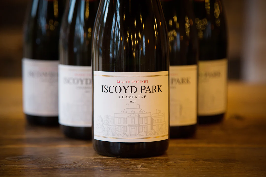Six bottles of Iscoyd Park Champagne with a gold label sit on a wooden table against an exposed brick wall background