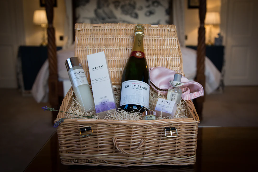 A hamper sits opened up displaying the contents which include a bottle of Neom bubble bath, a bottle of Iscoyd Park champagne, a neom candle and a pink silk eye mask