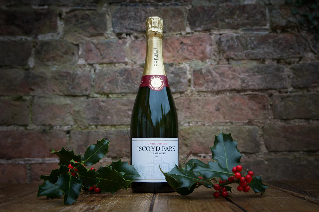 A single bottle of Iscoyd Park Champagne with a gold label surrounded by sprigs of holly sits on a wooden table against an exposed brick wall background