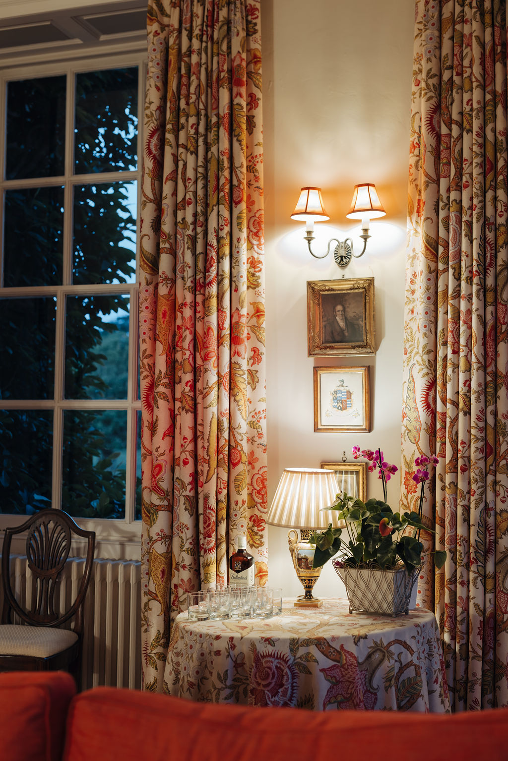 A vignette of a small drawing room at dusk with floor length floral curtains open at a large set of Georgian sash windows. In front of the window is a side table covered in a matching tablcoth on which is a vase of orchids and a lit table lamp