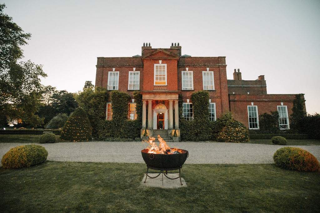A landscape view of a handsome red brick mansion with a grand stone portico with a fire pit burning in the foreground at dusk