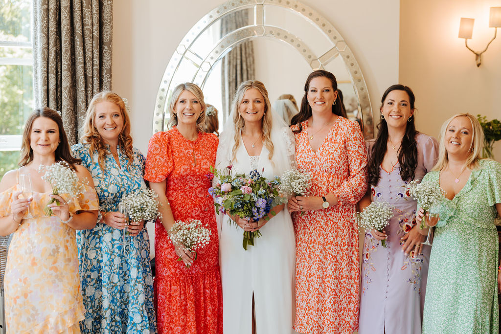Six adult bridesmaids wearing mismatched dresses in yellow, lilac, red, blue and green surround a bride whilst standing in front of a large wall hung oval mirror and holding their flower bouquets