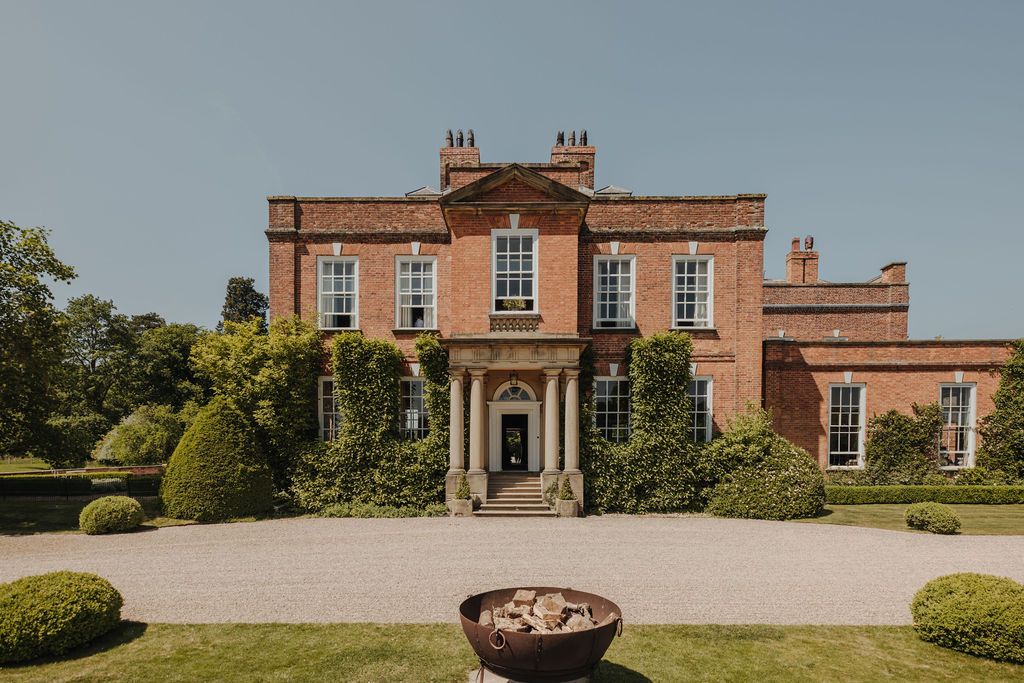A view of a handsome red brick mansion with a Portland Stone portico and landscaped gardens under a clear blue sky.