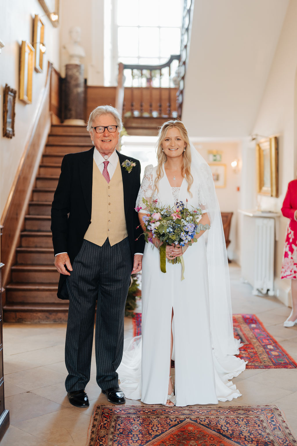 A man wearing a morning suit stands next to his daughter who is wearing a white lace wedding dress and holding a pastel coloured bouquet with an antique wooden staircase in the background behind them.