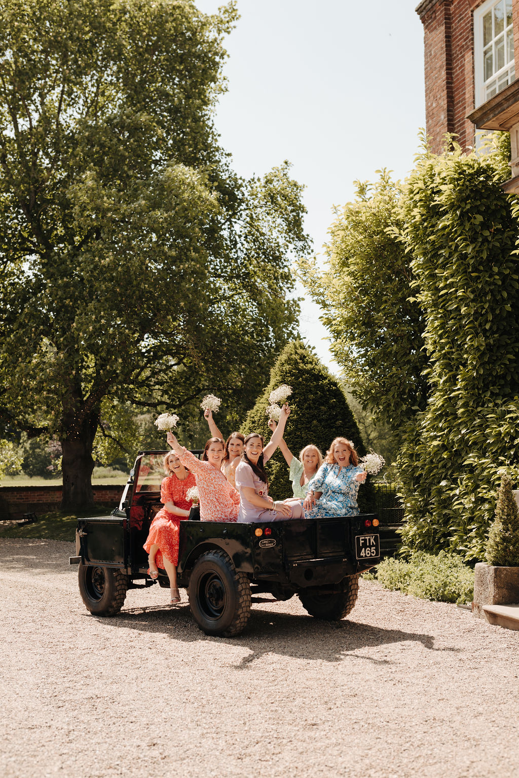 Six adult bridesmaids wearing mismatched dresses in yellow, lilac, red, blue and green sit in the back of an open top jeep waving their gypsophila bouquets aloft
