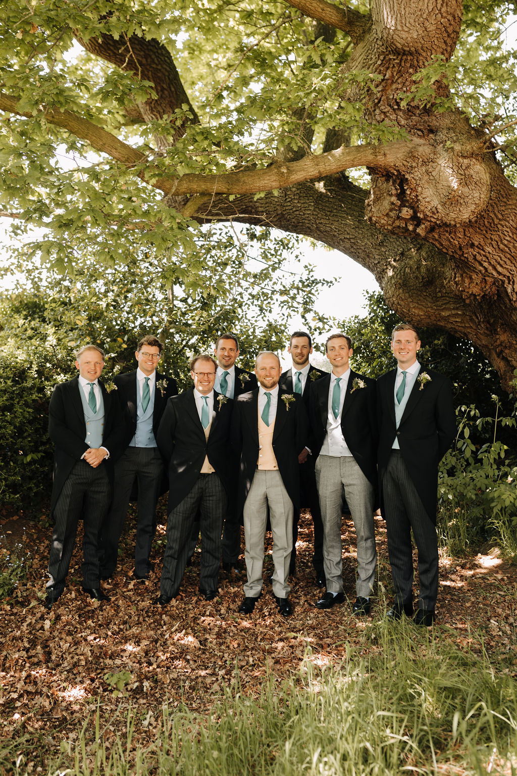 Eight adult men wearing morning dress and green ties for a wedding pose for the camera outside under a huge oak tree