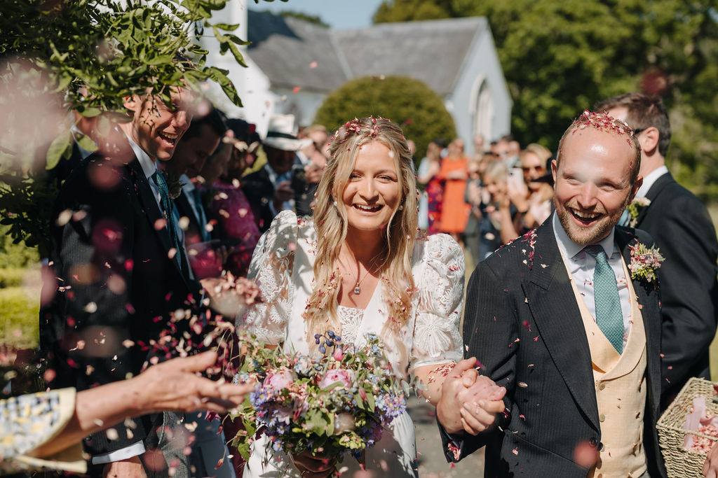 A bride and groom holding hands and smiling walk through guests throwing confetti with a white church in the background