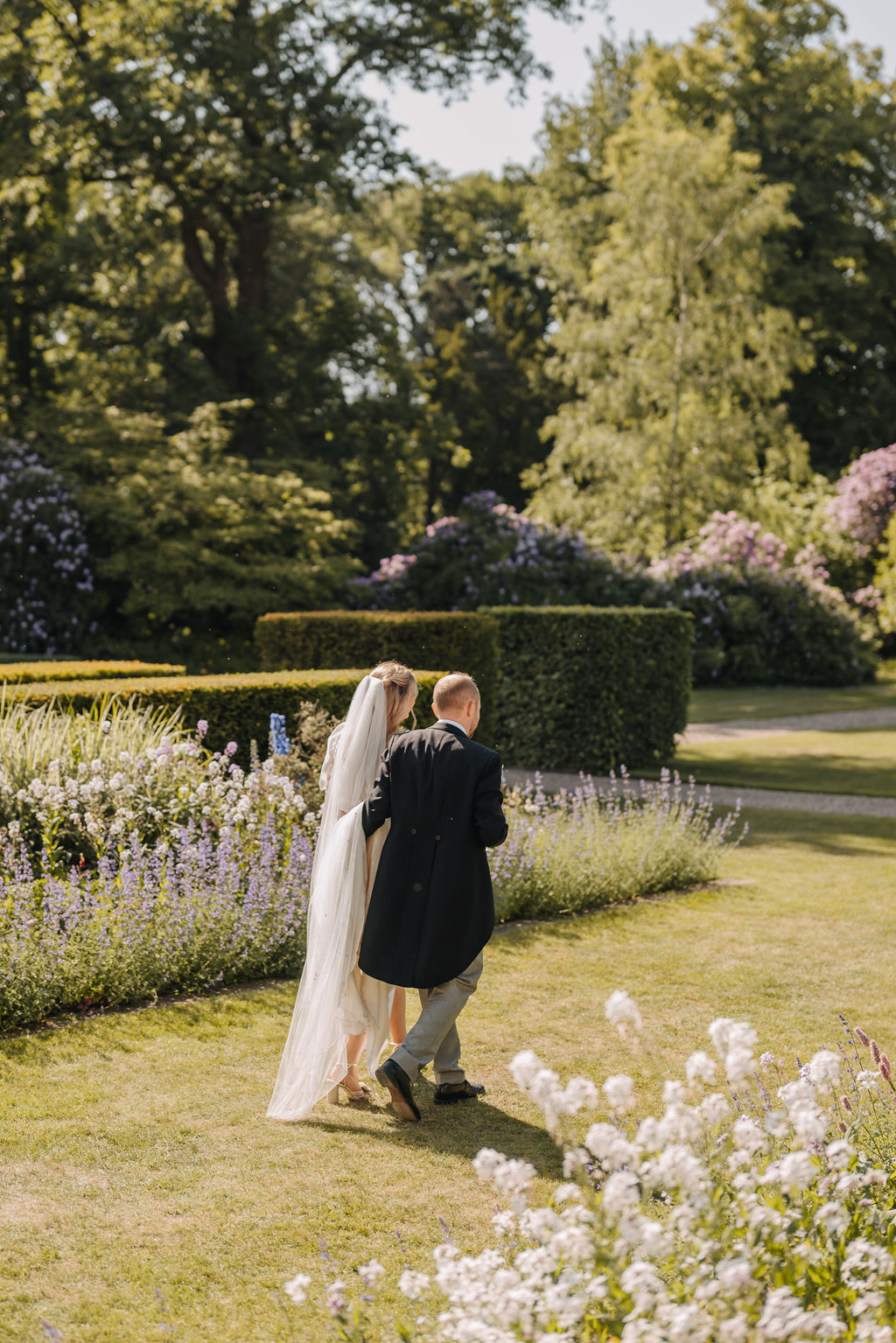 A bride and groom walk along a green lawn flanked by established garden beds filled with summer flowers