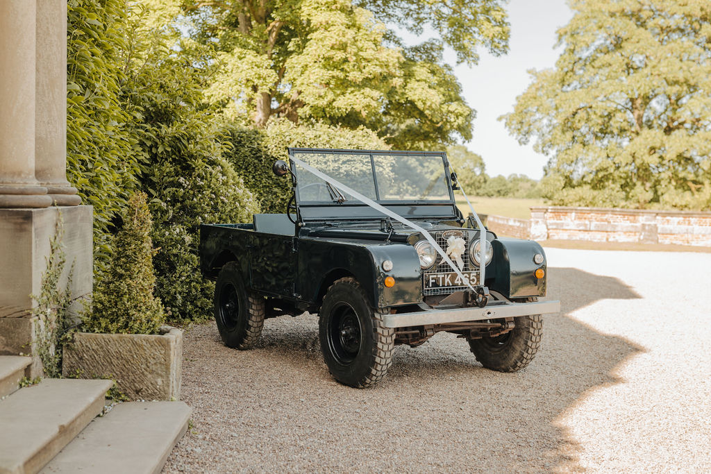 An old green military jeep is parked outside a red brick mansion covered in wedding ribbons