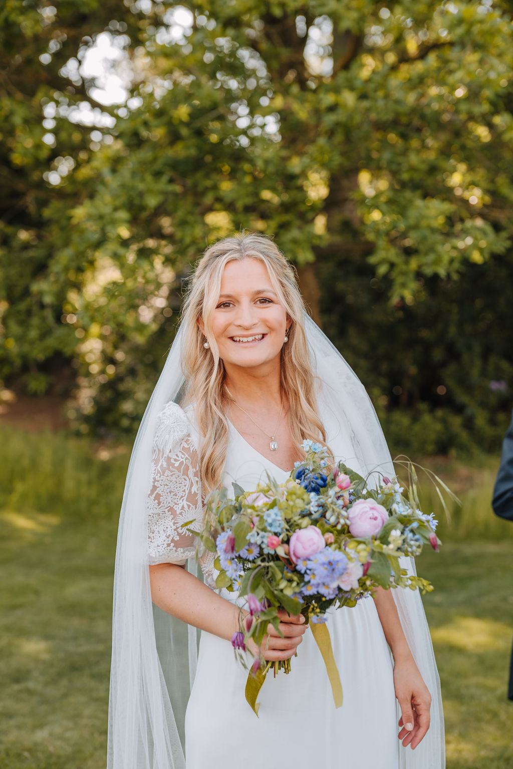 A bride wearing a white wedding dress with lace sleeves and a pastel toned bouquet wears her hair down under a long tulle veil and smiles at the camera