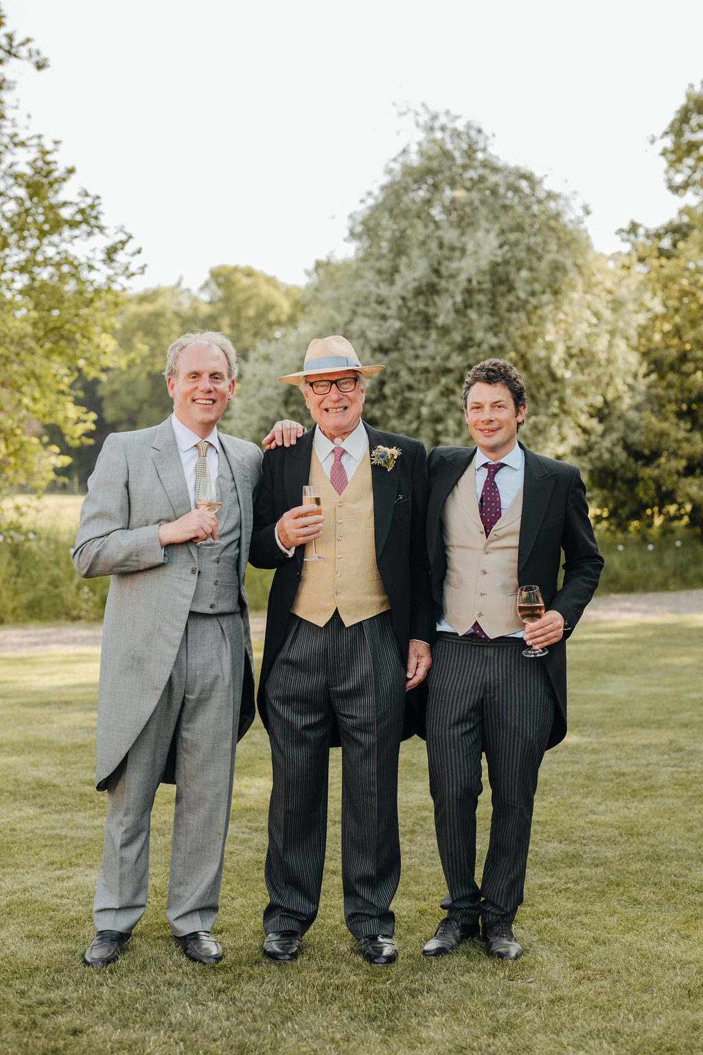 Three men wearing formal three piece suits stand and pose for the camera in a beautiful garden whilst holding glasses of champagne