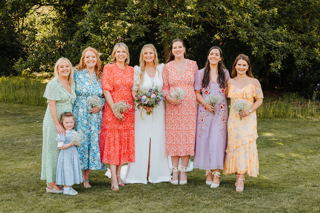 Six adult bridesmaids wearing mismatched dresses in yellow, lilac, red, blue and green surround a bride and a little flower girl wearing a blue smocked dress whilst standing outside on a grassed areas
