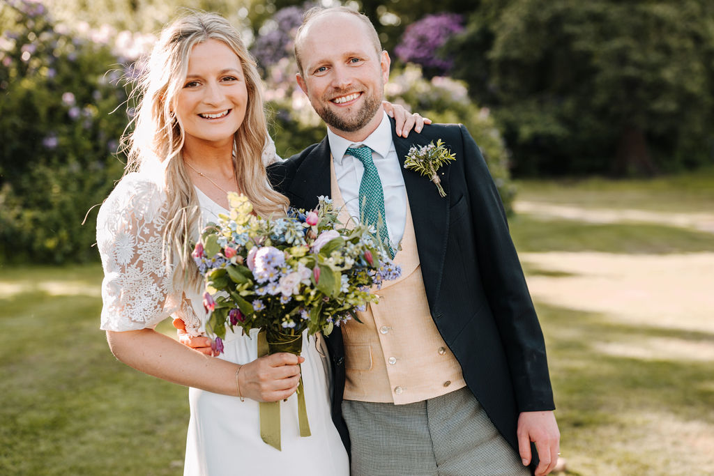 A bride clasps her husband around the shoulders as the groom wears a morning suit and the bride wears a long dress with lace sleeves holding a bouquet of pastel wild flowers.