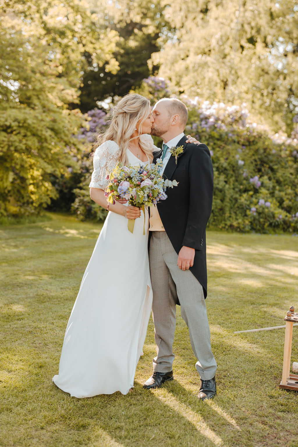 A bride and groom kiss as the groom wears a morning suit and the bride wears a long dress with lace sleeves holding a bouquet of pastel wild flowers.