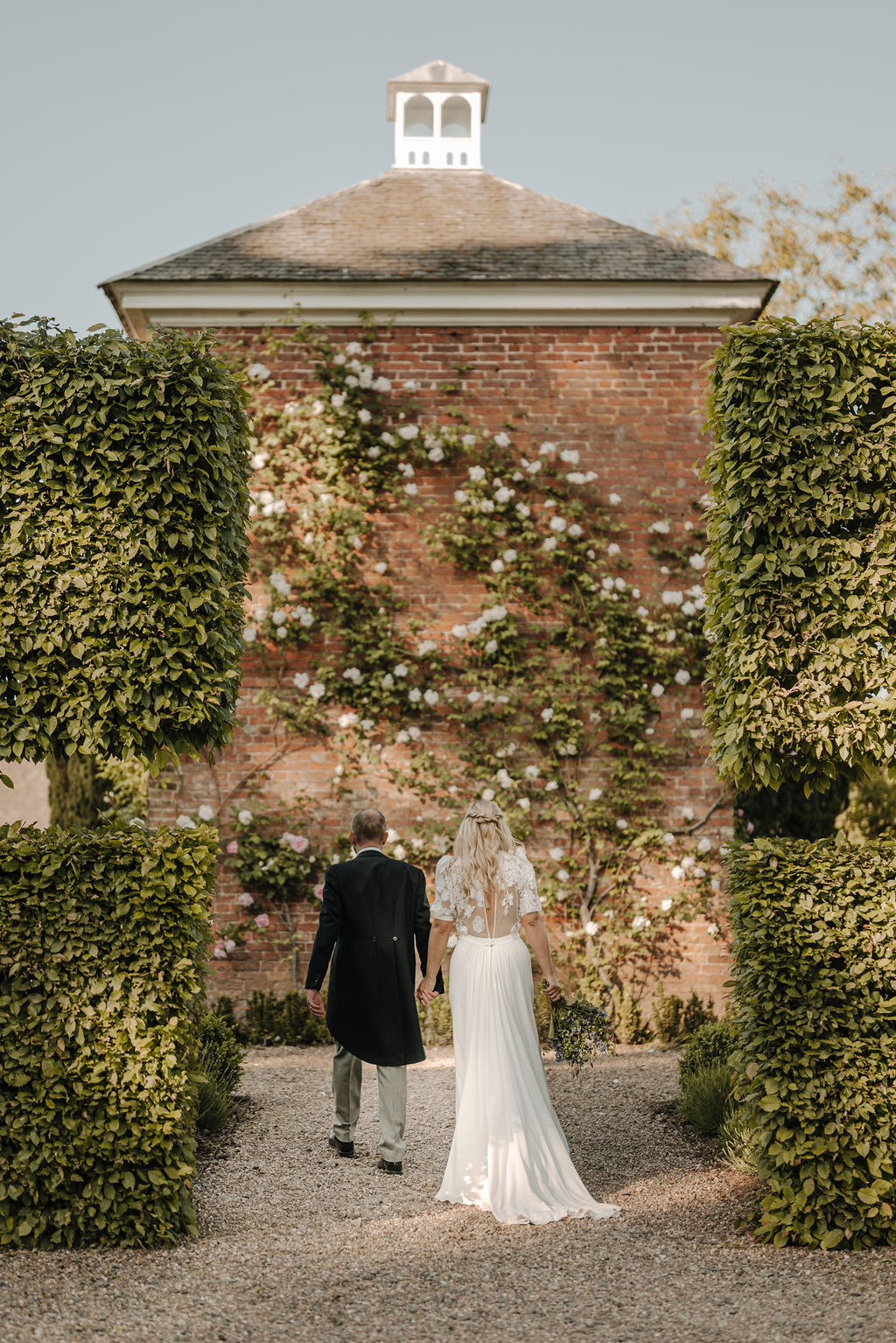 A bride and groom walk away from the camera along a gravel path with a red brick building in the background covered with a white climbing rose