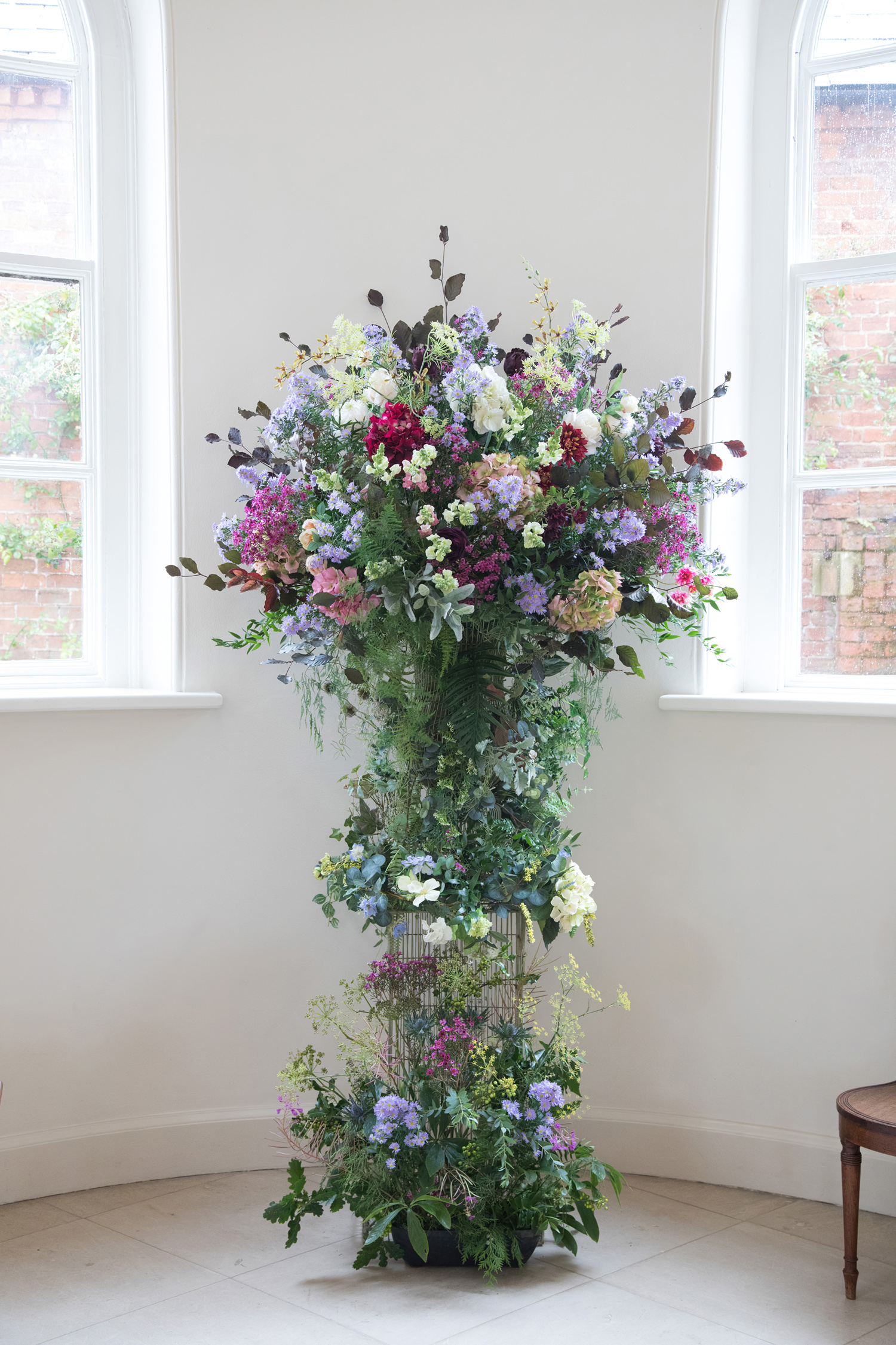 A focused image on a large floor standing floral arrangement for a wedding using blue, white, lilacs and deep red colour schemes and lots of green foliage