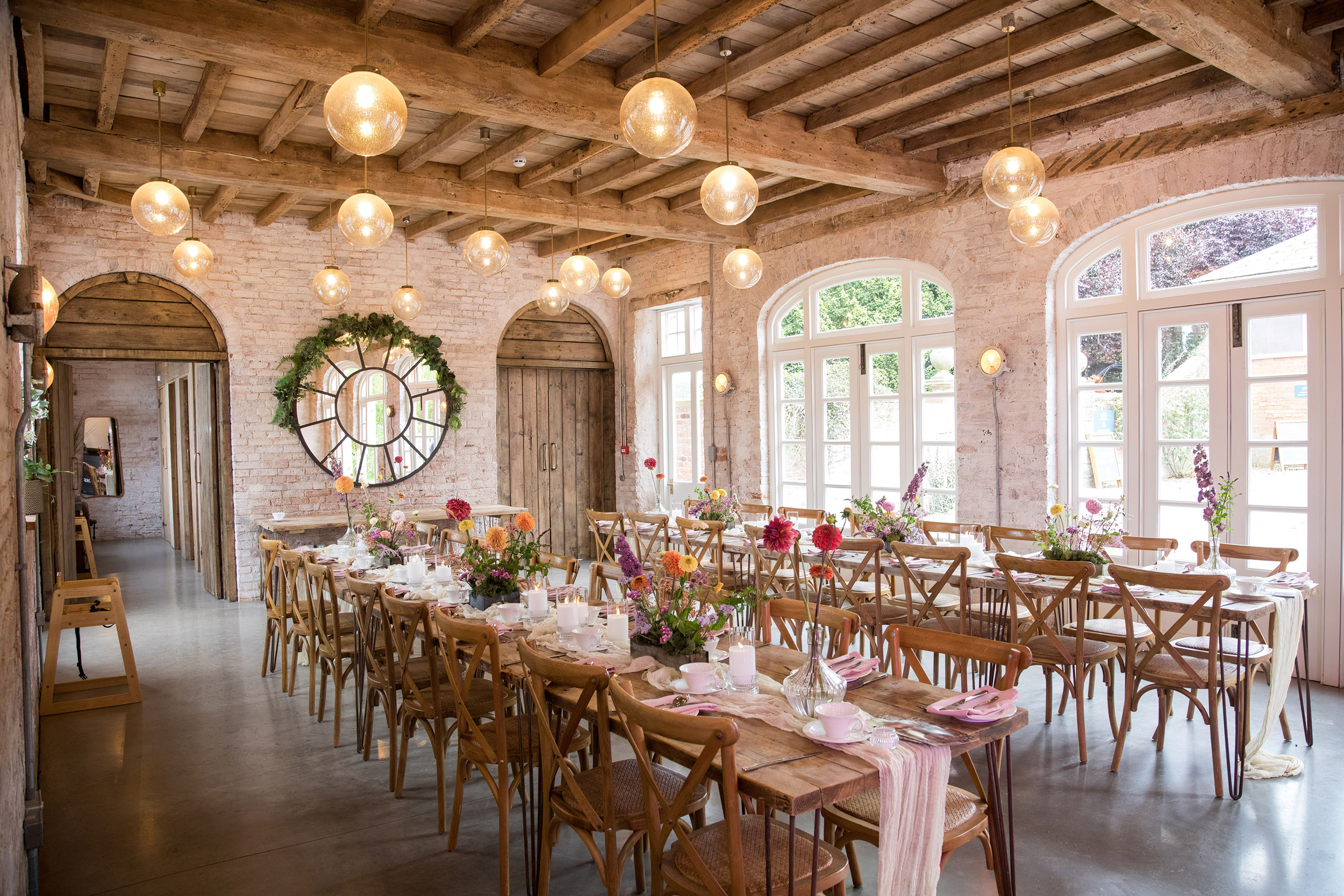 A view of two rows of trestle tables laid out for a wedding reception in a rustic exposed brick wall room with exposed beams. The tables have small bud vases filled with single stems of dahlias in reds, oranges and yellows