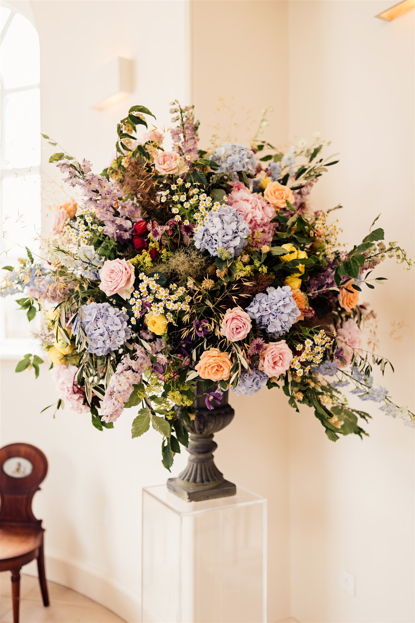 A large urn filled with floral arrangements filled with green foliage and pink, blue and lilac tones set on a plinth in a neutral room