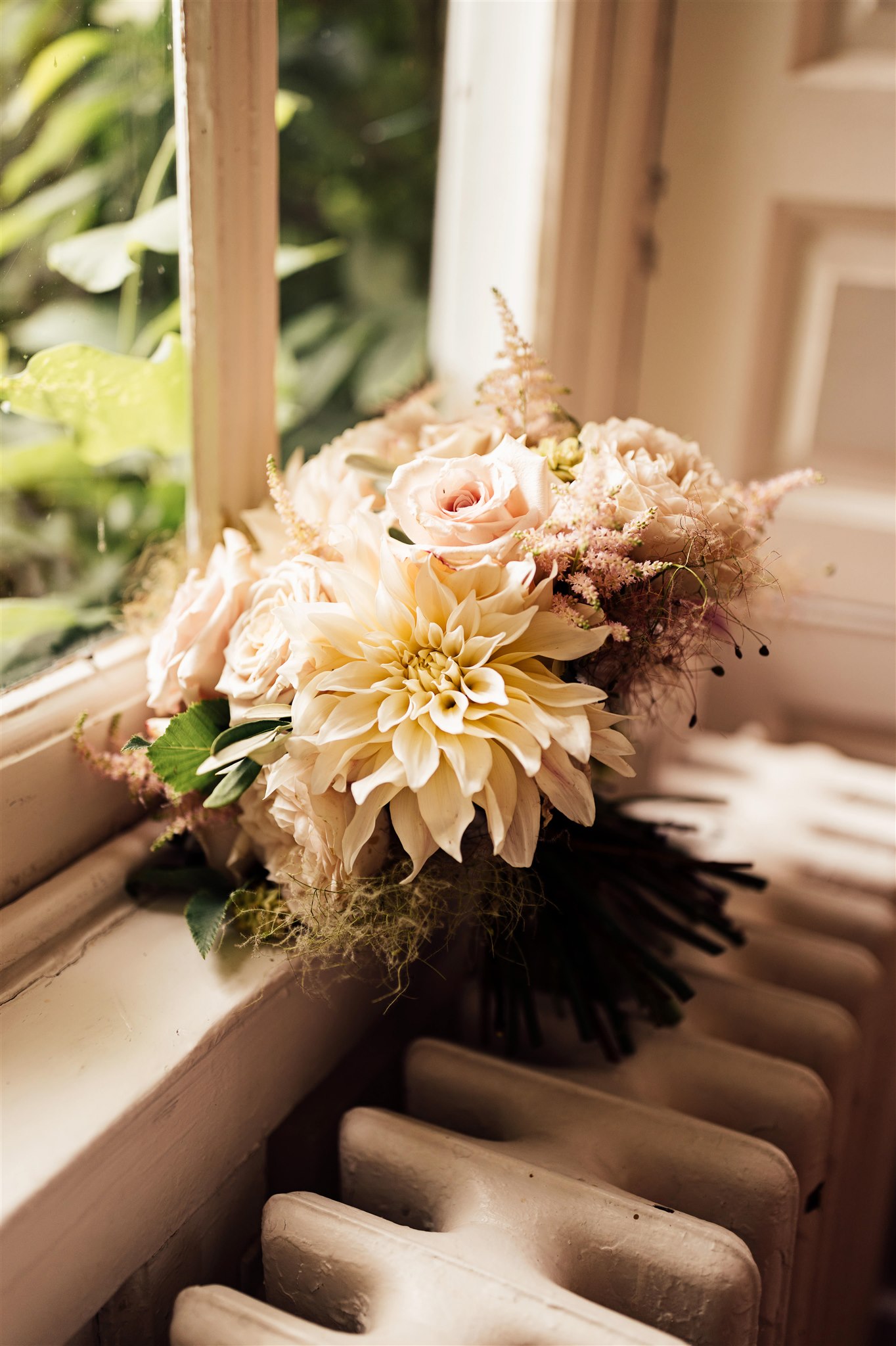 A wedding bouquet made up of roses and dahlias in blush pink and caramel tones sits on a radiator next to a window