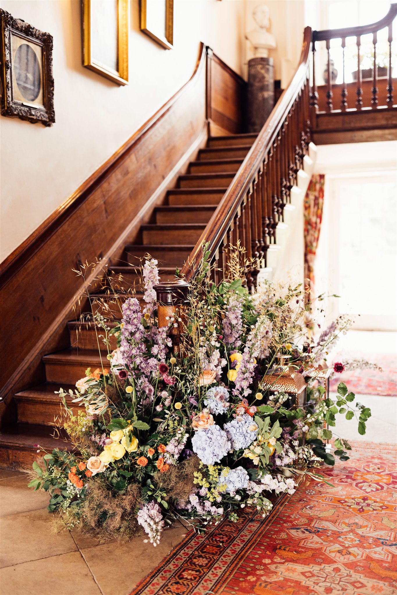 A floor floral arrangement filled with green foliage and pink, blue and lilac tones sits next to an old wooden staircase