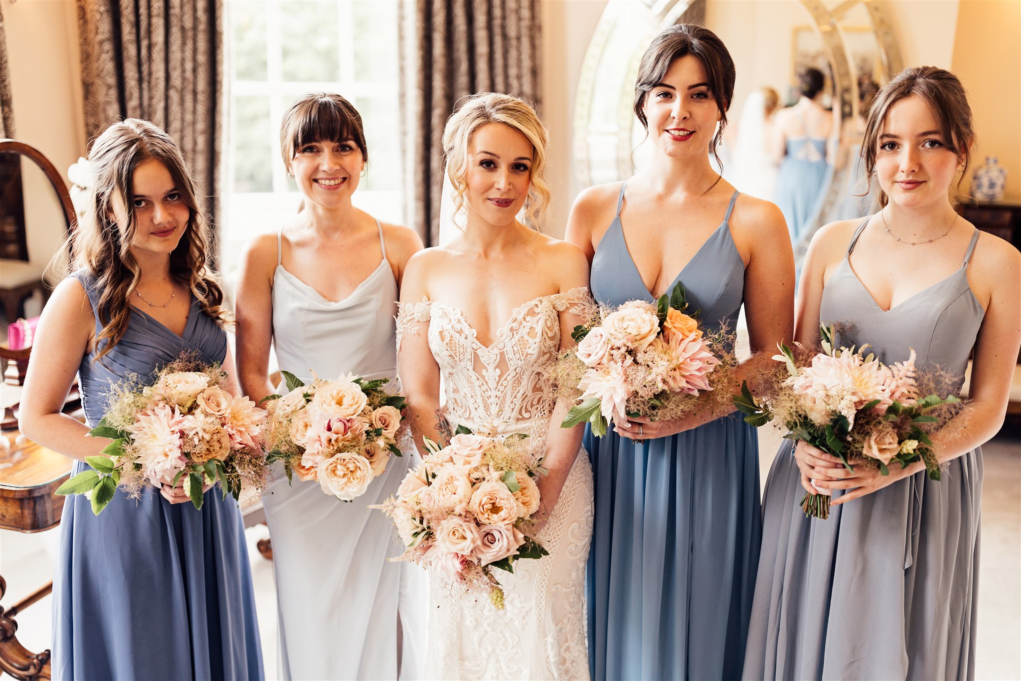 A bride wearing an off the shoulder white lace dress with blonde hair stands with two bridesmaids on either side of her. The bridesmaids wear blue floor length dresses and all five women are holding blush pink bouquets made up of dahlias and roses.