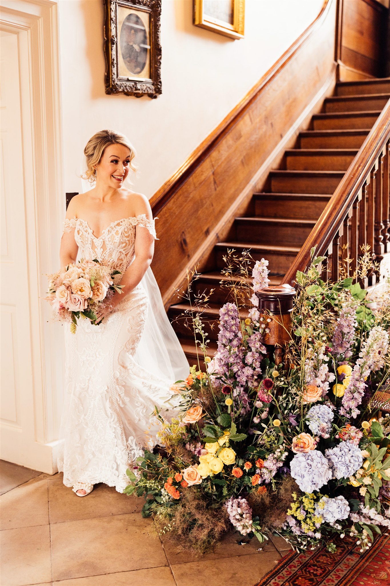 A floor floral arrangement filled with green foliage and pink, blue and lilac tones sits next to an old wooden staircase down which a bride is descending