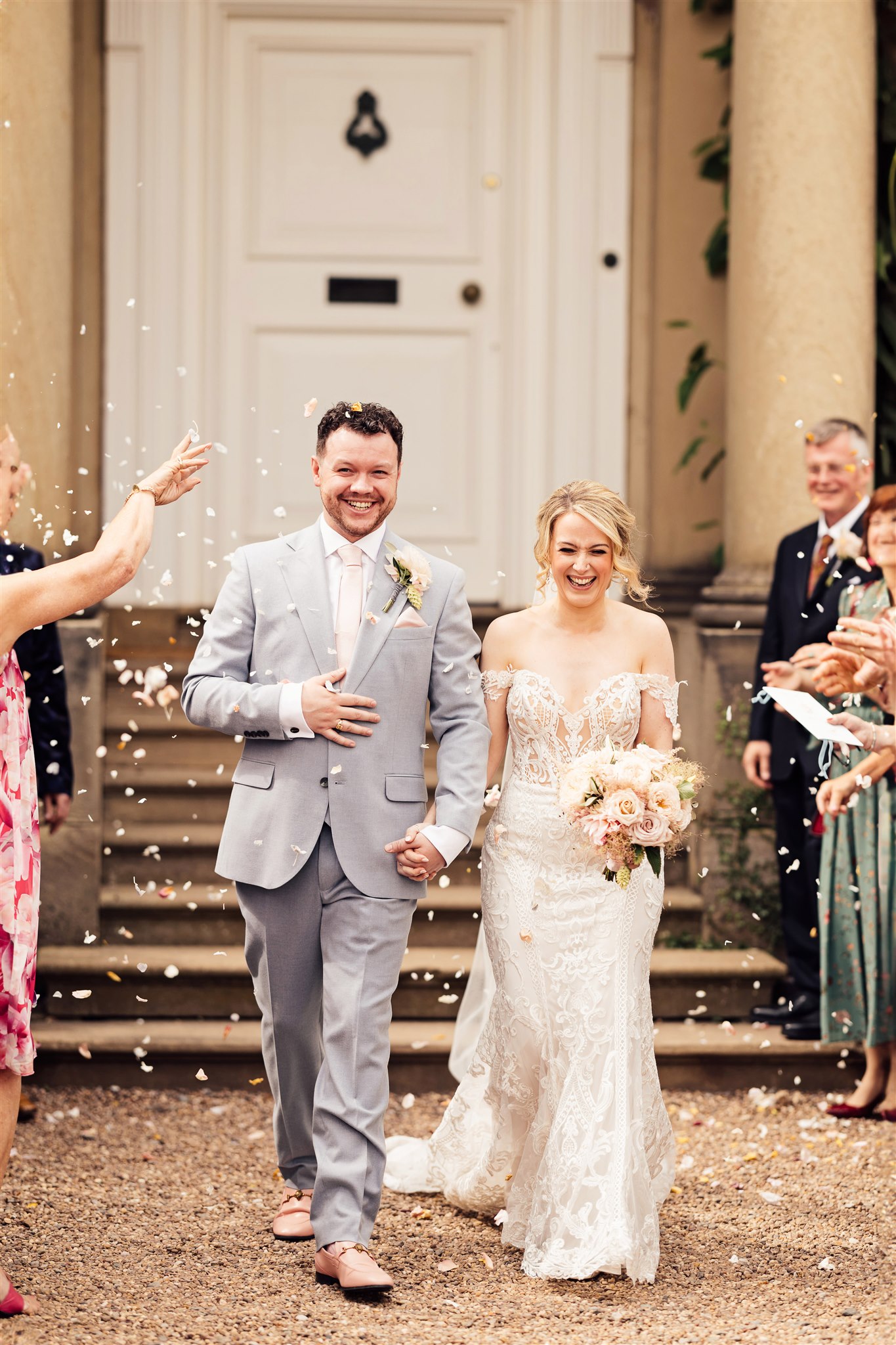 A bride wearing an off the shoulder white lace dress stands next to a groom wearing a grey suit and pink tie with the background of a red brick Georgian mansion and their guests throwing confetti over them