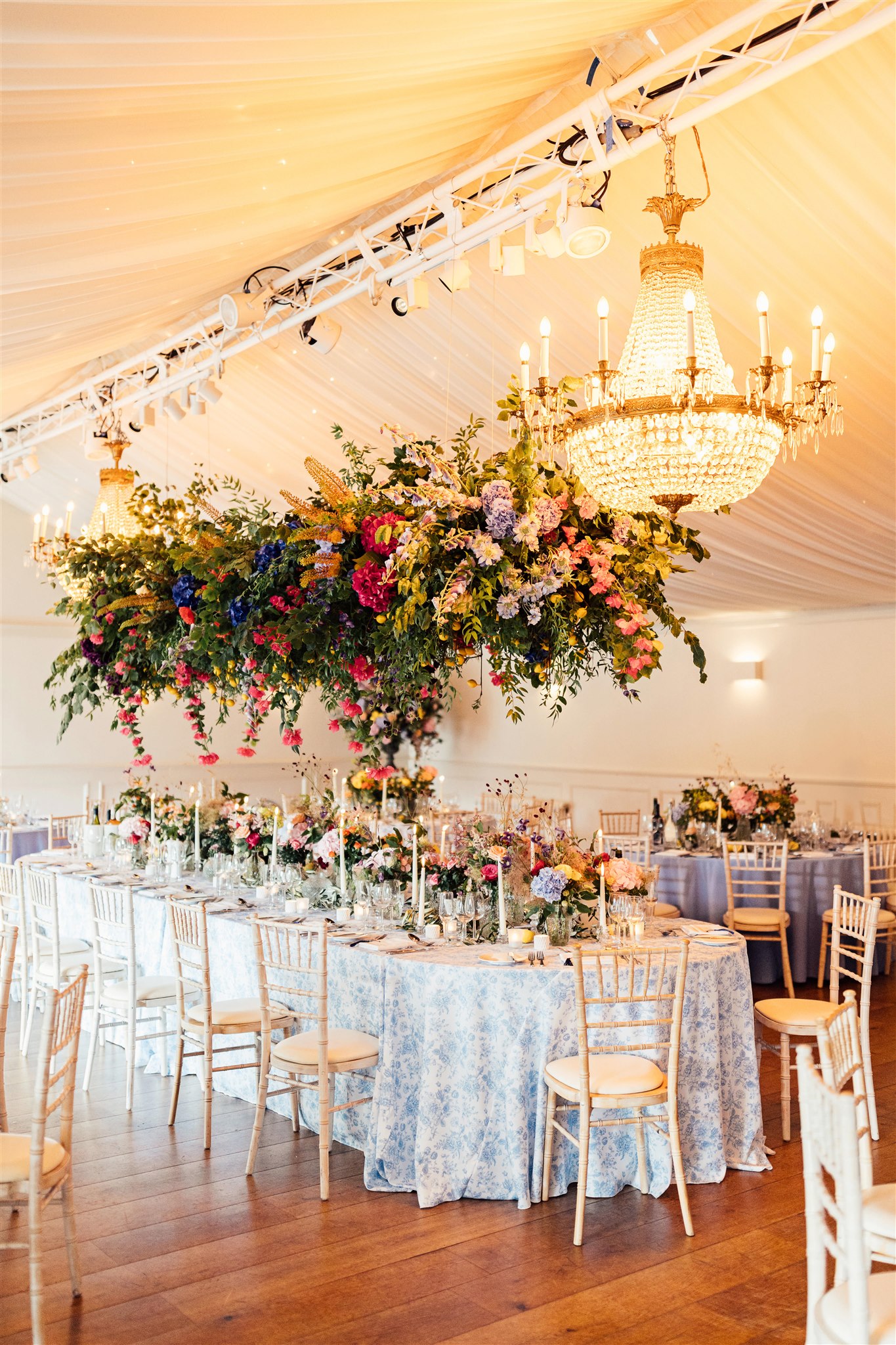 A huge crystal chandelier is suspended from the ceiling of a wedding marquee next to a hanging floral arrangement filled with green foliage and pink, blue and lilac tones. Underneath is an oval table with a blue toile tablecloth on which are more flowers and cream lit candles.