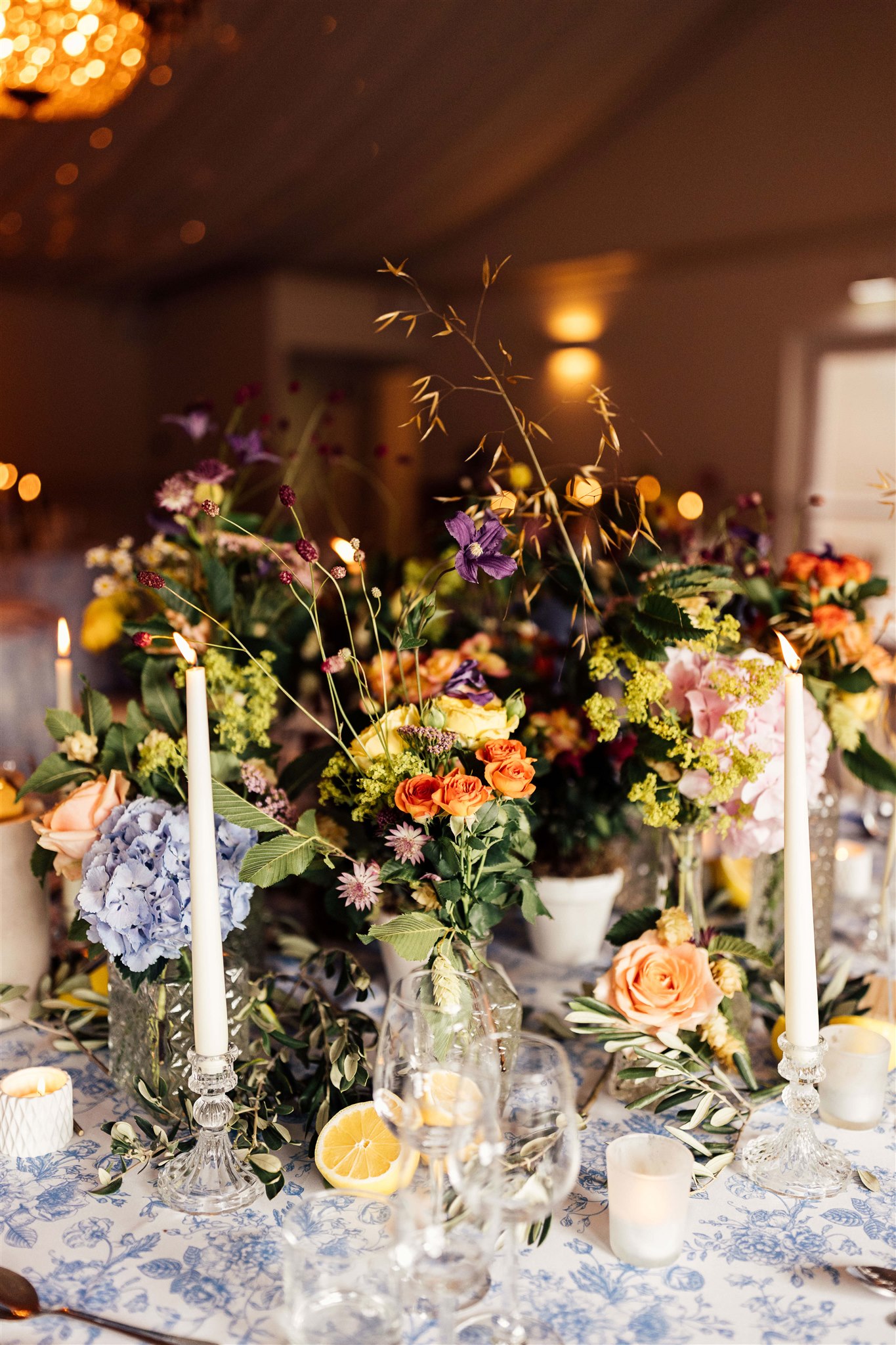 A table filled with floral arrangements filled with green foliage and pink, blue and lilac tones set on a blue toile tablecloth on which are more flowers and cream lit candles.