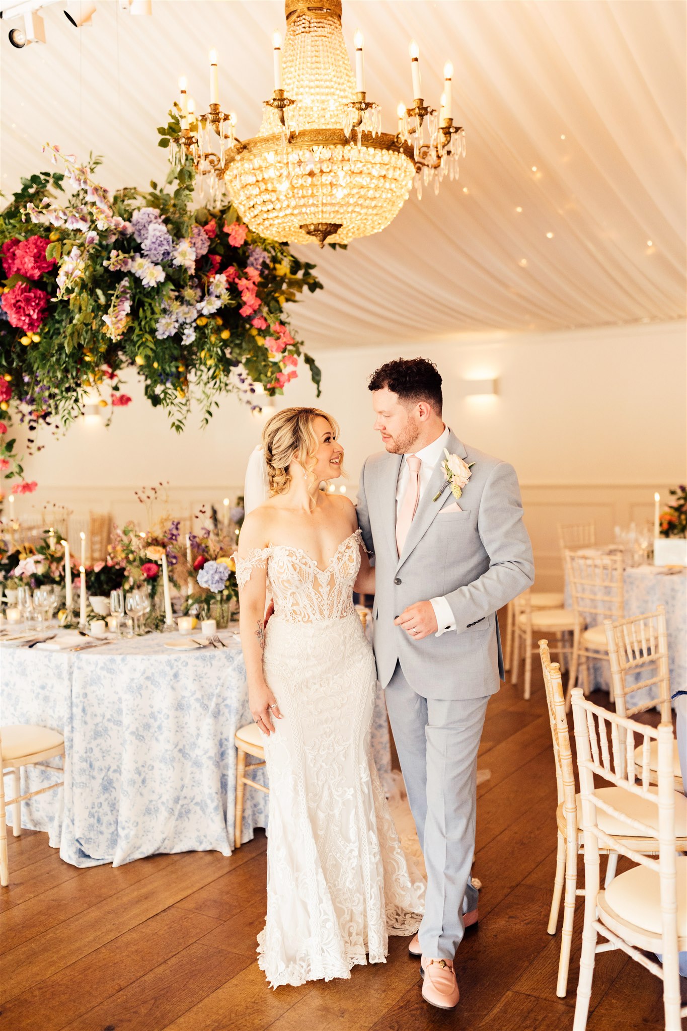 A huge crystal chandelier is suspended from the ceiling of a wedding marquee next to a hanging floral arrangement filled with green foliage and pink, blue and lilac tones. Underneath is an oval table with a blue toile tablecloth on which are more flowers and cream lit candles.