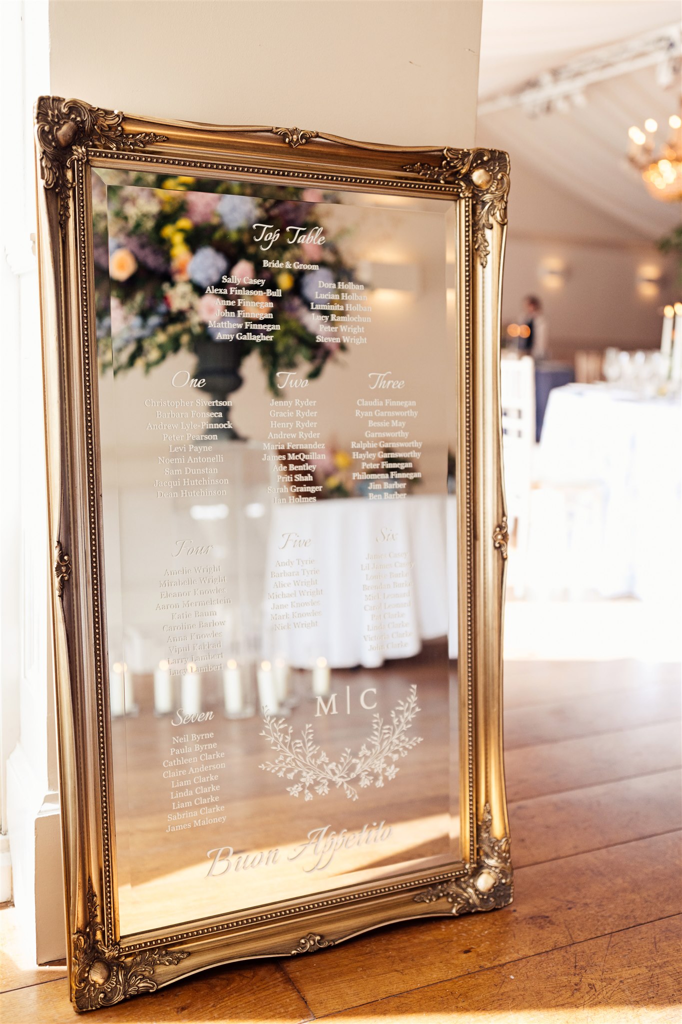 A huge mirror with a gilt gold frame stands on the floor in the entrance of a wedding marquee and is inscribed with a wedding table plan written in white script on the glass of the mirror itself
