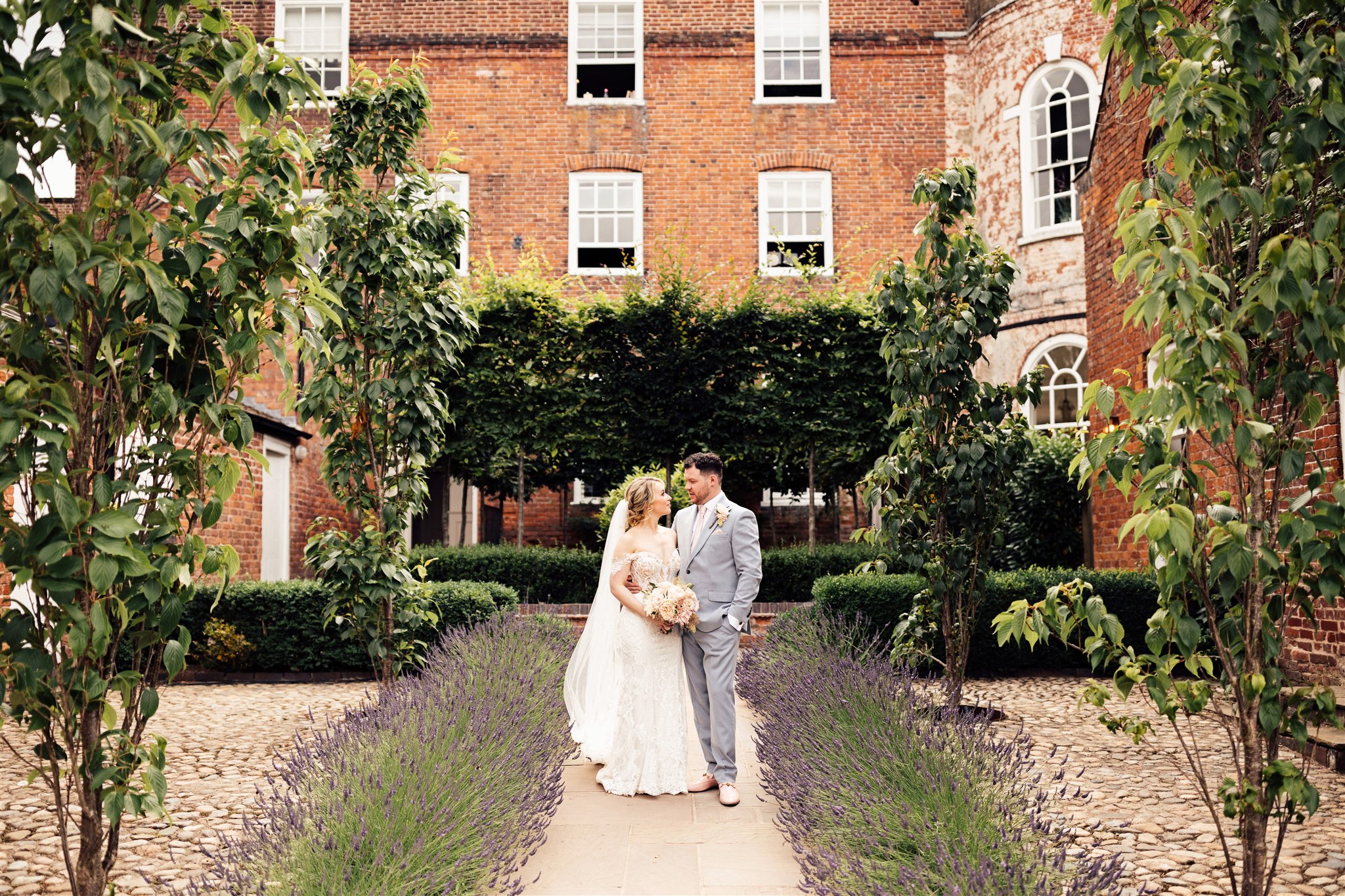 A bride wearing an off the shoulder white lace dress stands next to a groom wearing a grey suit and pink tie with the background of a red brick Georgian mansion with an alley of lavender in the foreground