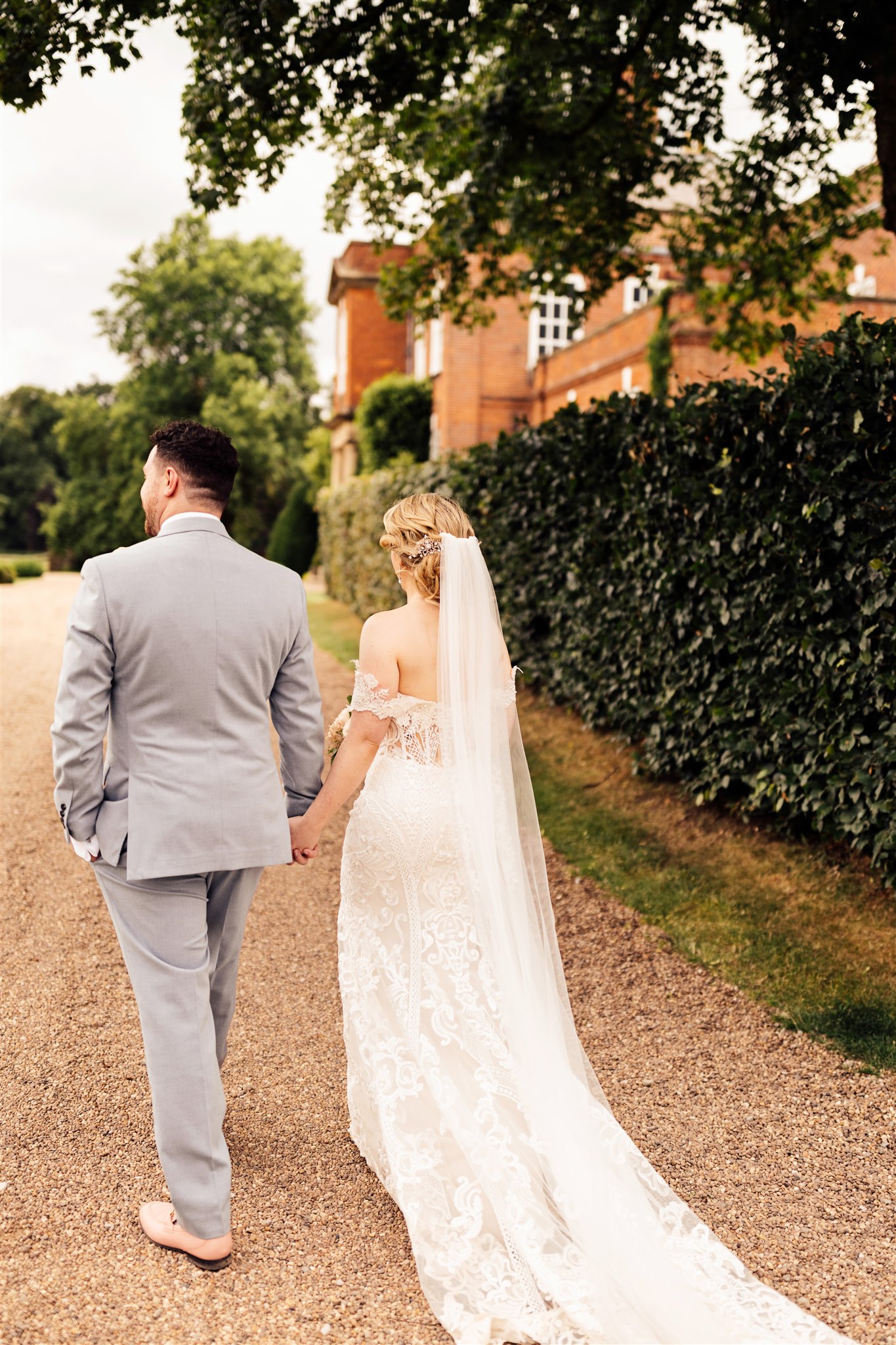 A bride wearing an off the shoulder white lace dress stands next to a groom wearing a grey suit and pink tie with the background of a red brick Georgian mansion.