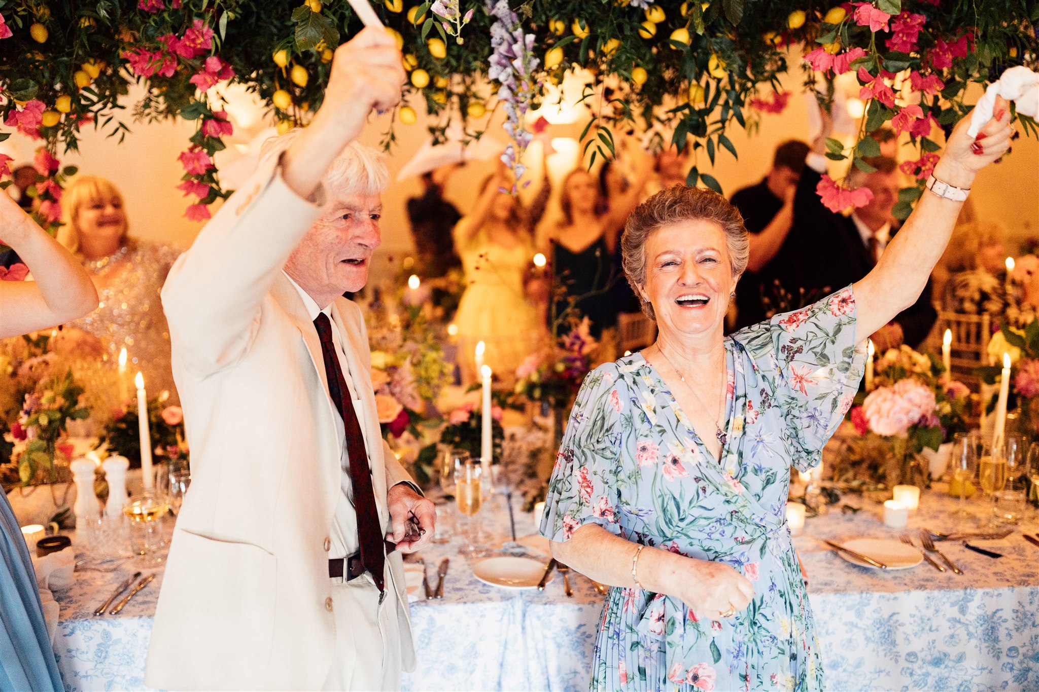 A middle aged lady wearing a floral dress stands waving a napkin and smiling next to an elderly man wearing a beige suit. They are celebrating at a wedding