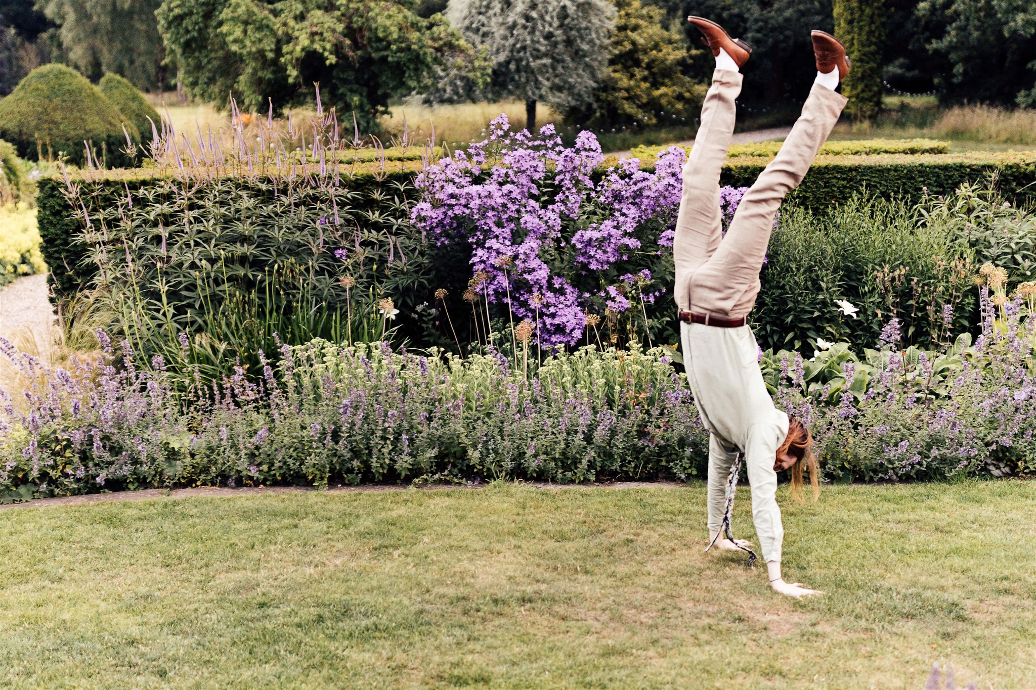 A man with beige trousers and a cream shirt and floral tie is captured doing a handstand in an English country garden against a backdrop of catmint and salvias
