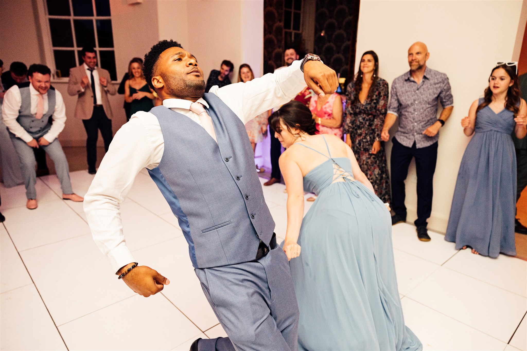 A man with a blue three piece suit and pink tie and a cream shirt is caught in a crazy dance move on the dance floor whilst other wedding guests look on.