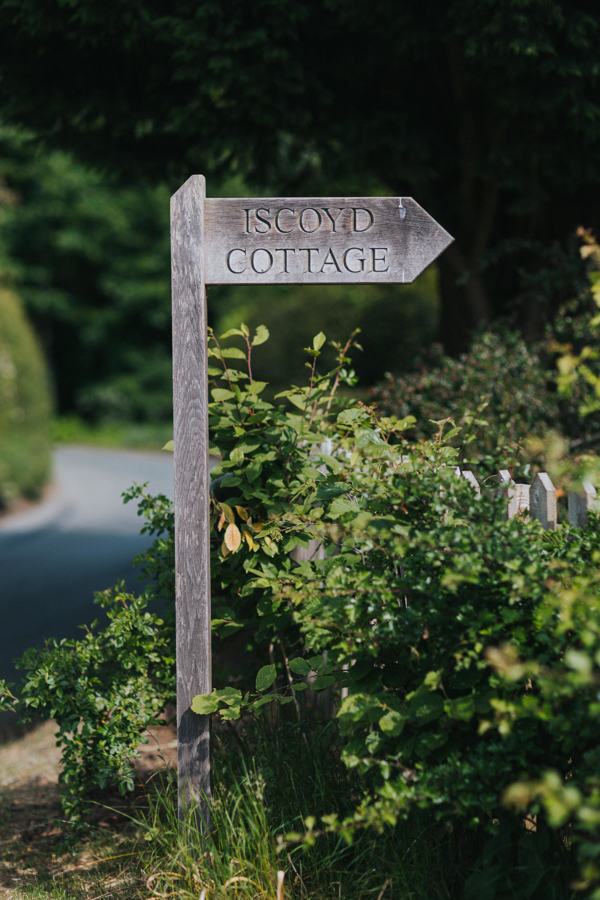 A wooden signpost which has been engraved with the words Iscoyd Cottage points to the right surrounded by native British hedgerows