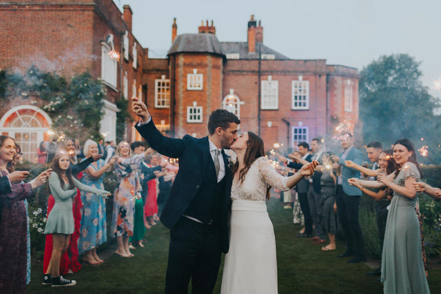 A bride and groom kiss whilst holding sparklers in their hands with their wedding guests surrounding them with a redbrick country manor stands in the background