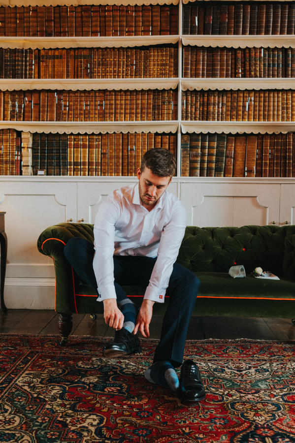 A groom sits on a sofa tying his smart black shiny wedding shoes with a backdrop of antique books on bookshelves behind him