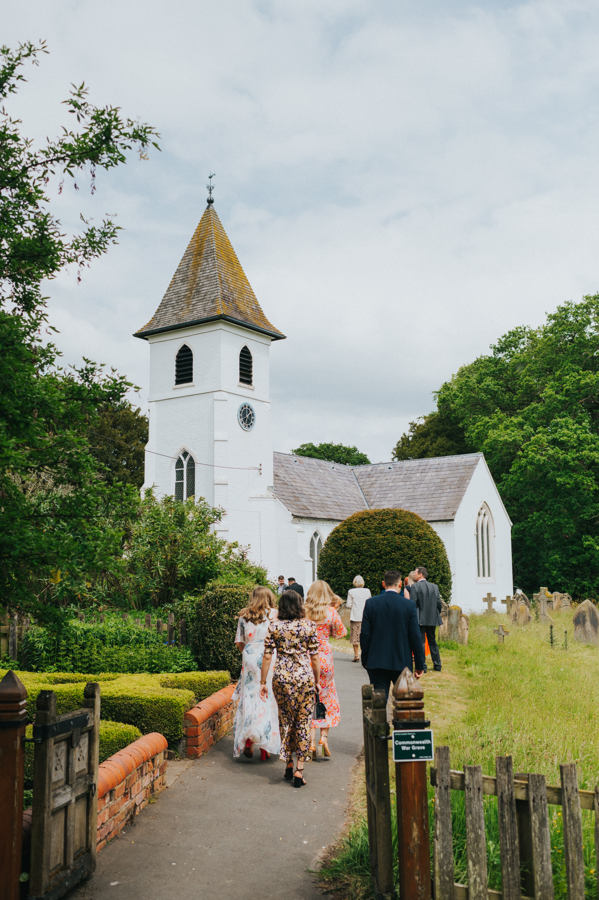 A view of a painted white church set amongst a countryside setting with wedding guests walking up a path towards the church