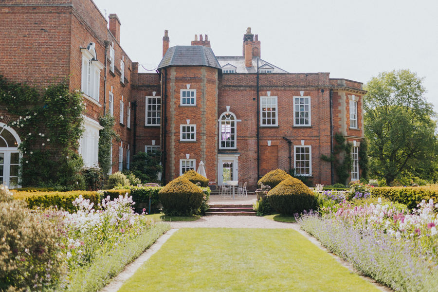 A view of an imposing redbrick Georgian mansion set in manicured gardens with flowerbeds of perennial English country garden flowers